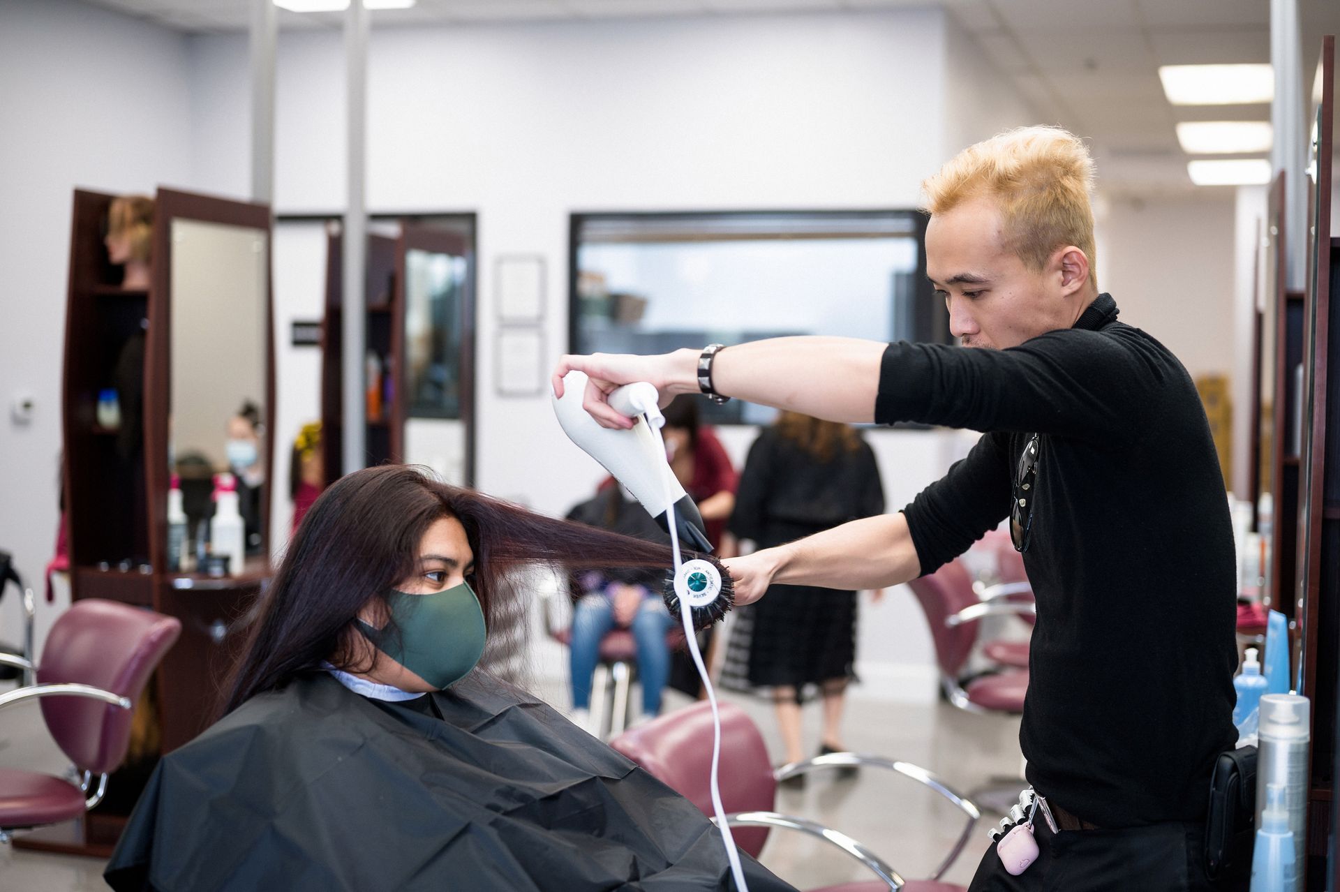 Hair stylist blow-drying client's hair in salon. Client wears mask and cape. Mirrors and other clients in background.