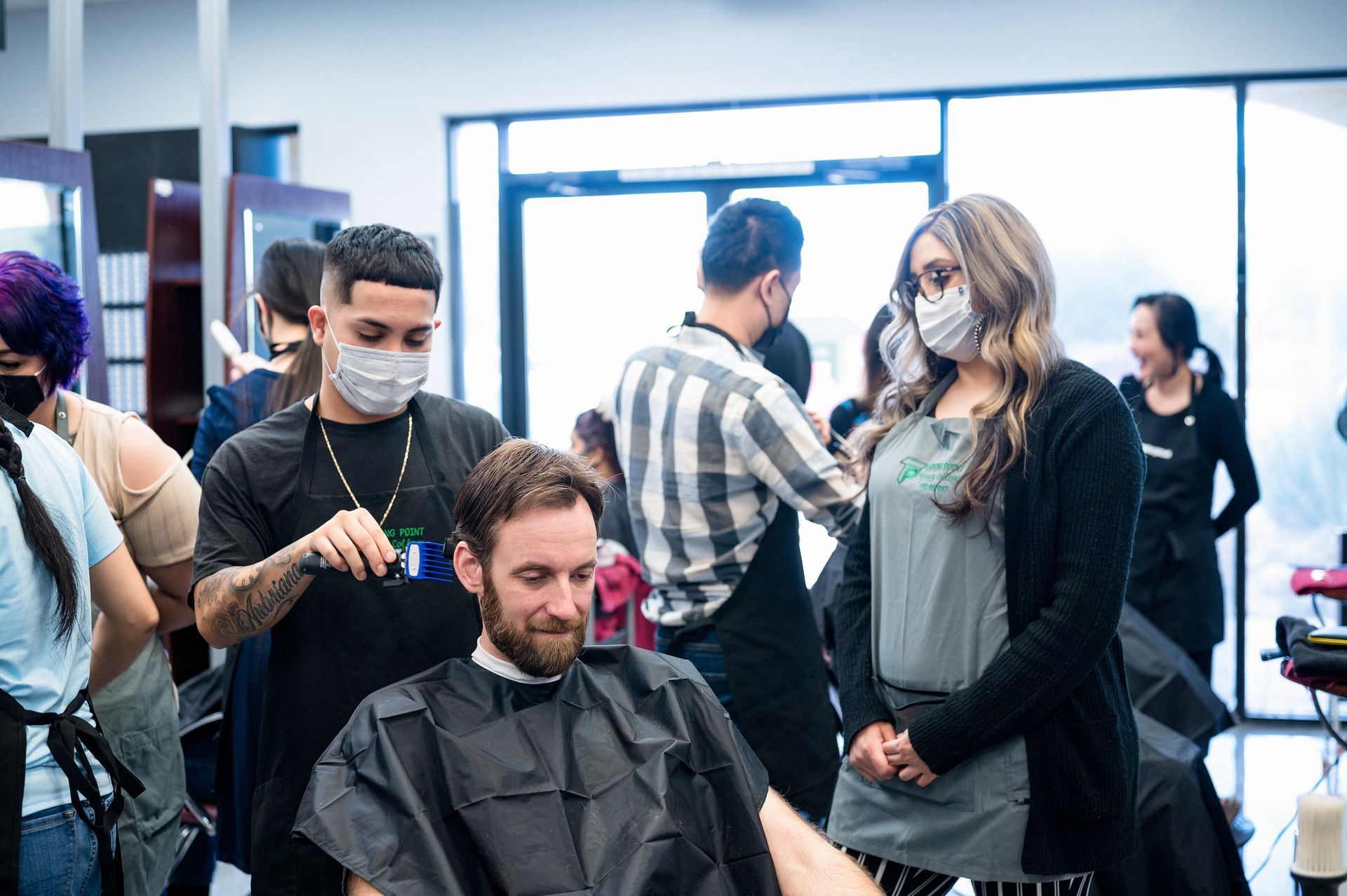 A barber giving a haircut to a client at a salon; several people, all wearing masks, are in the background.