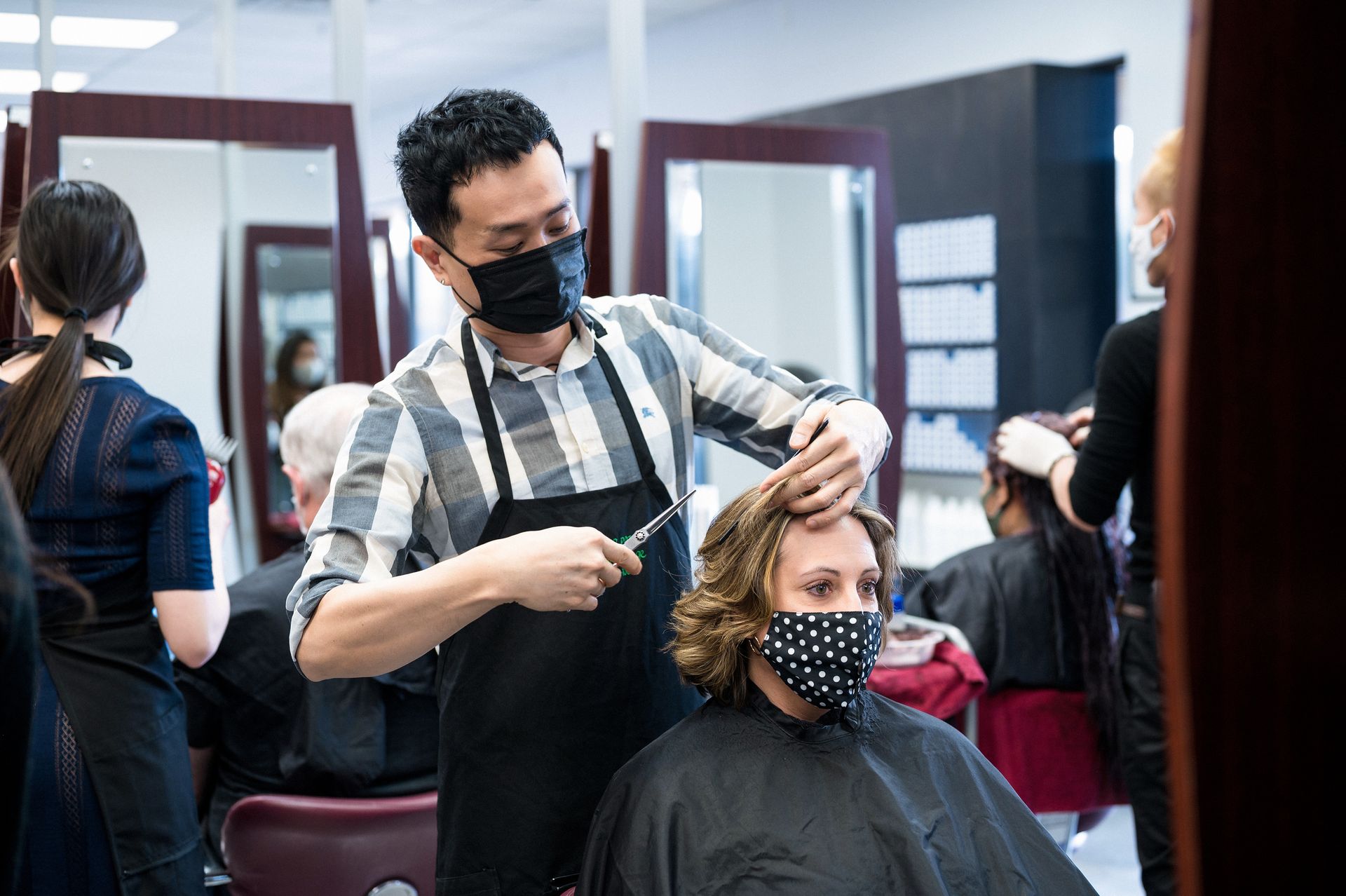 Man in mask cutting a woman's hair in a salon; both wear masks.
