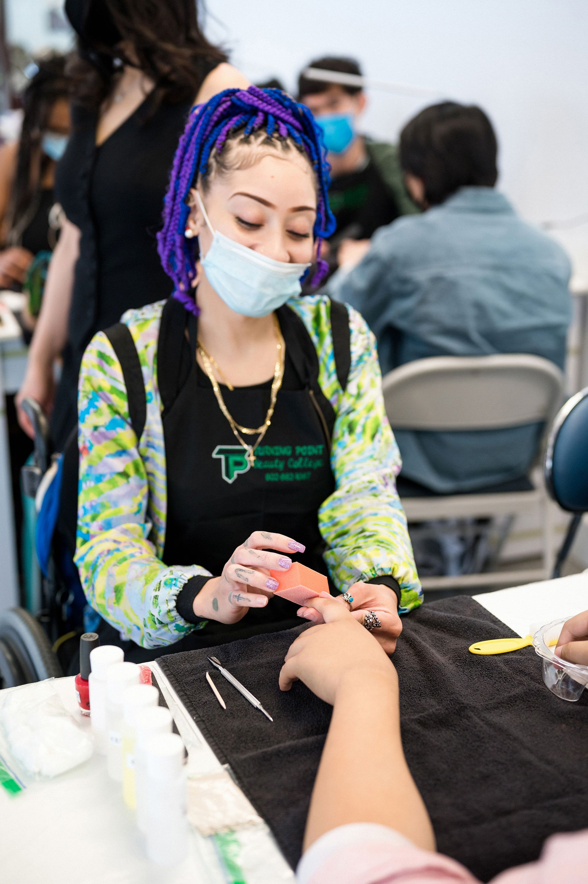 Woman with purple braids files a person's nails while wearing a mask and apron, indoors.