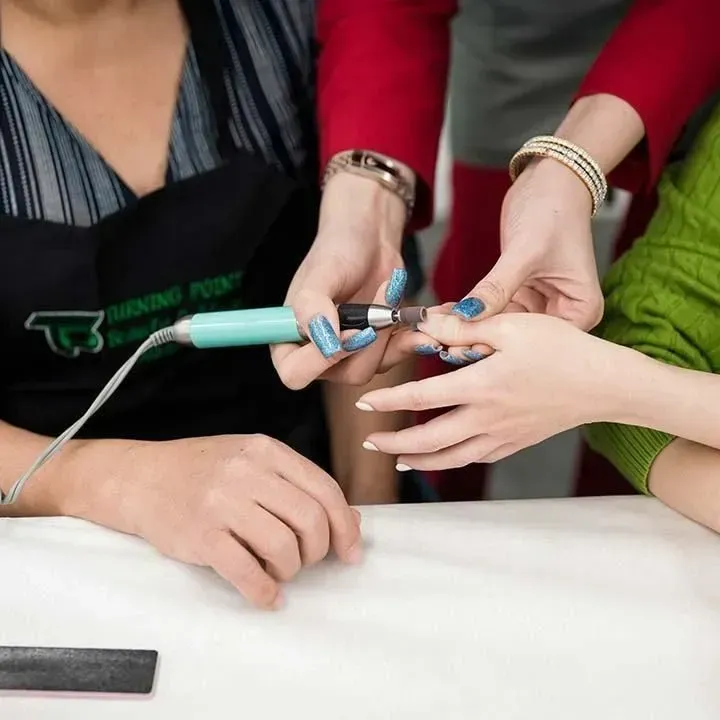 Hands using a nail drill on a person's fingernail at a table; another person assists.
