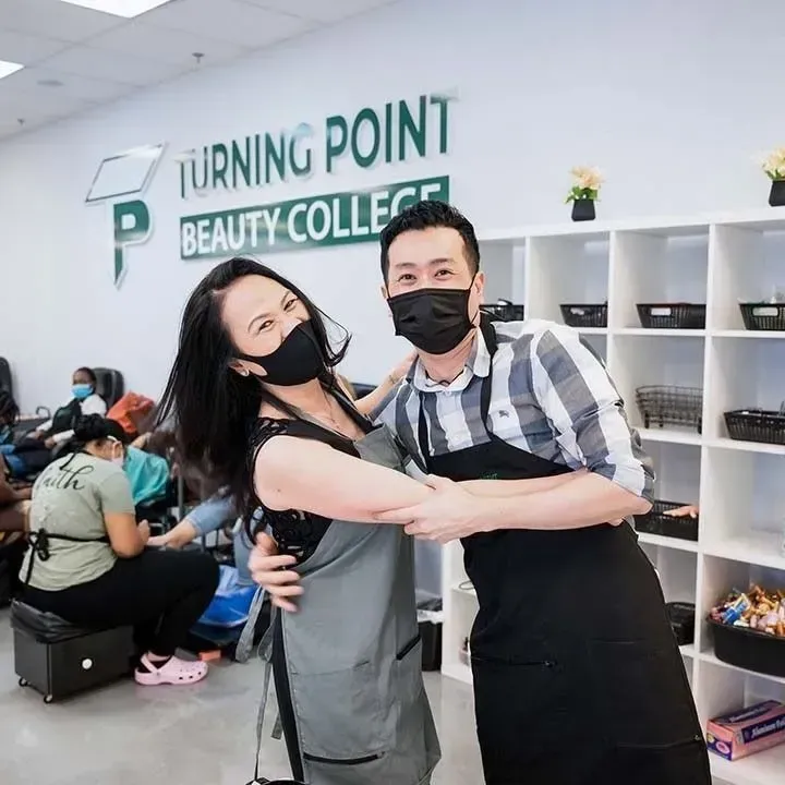 Two people wearing masks and aprons pose in a beauty college, smiling.