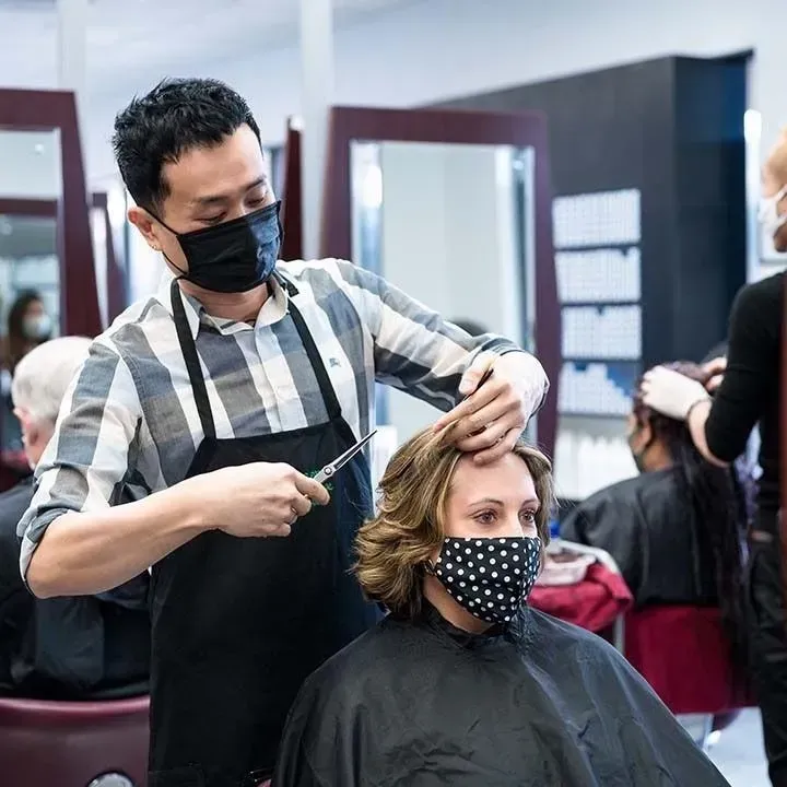 Hair stylist wearing a mask cuts a client's hair in a salon. Both are masked; other masked clients present.