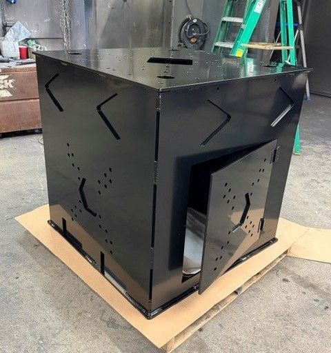 Black metal box with cutout vents, sitting on cardboard in a workshop or garage.