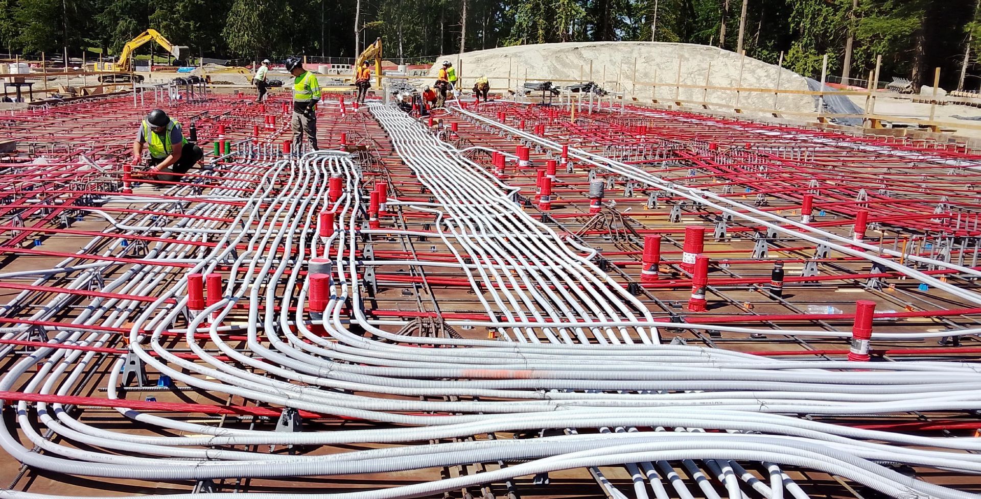 Construction site: Workers installing conduits on a foundation. White and red pipes, red supports.