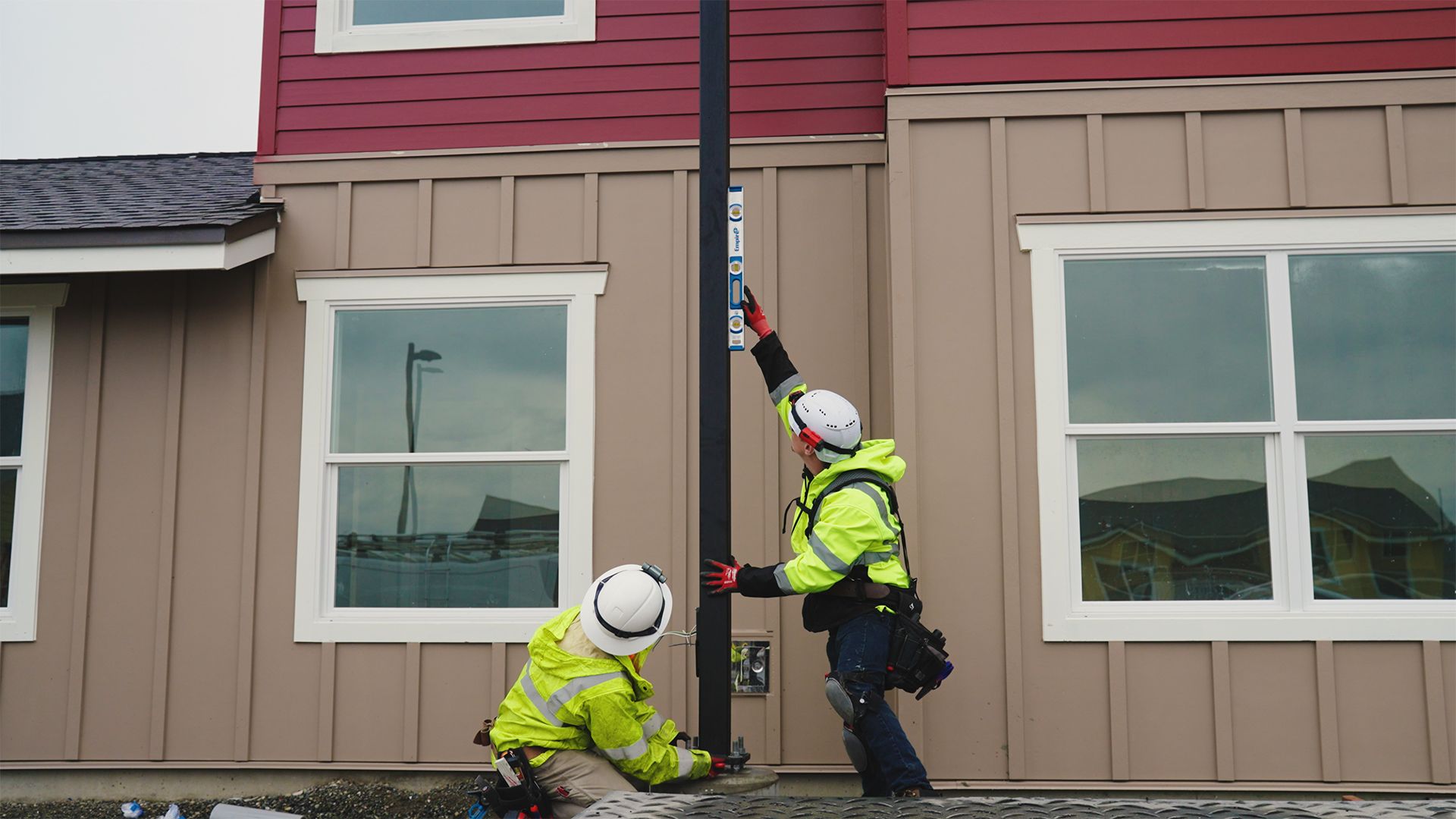 Two utility workers in safety vests installing a black pole on a house