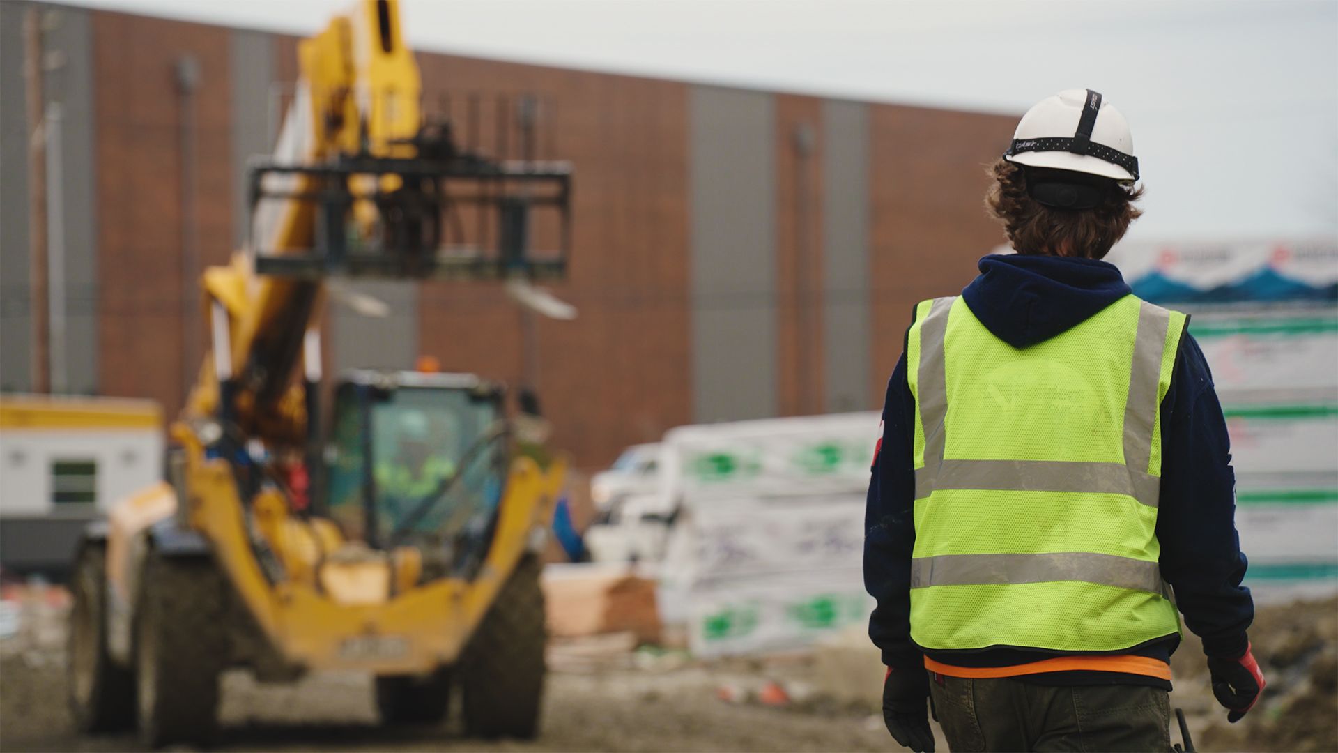 Construction worker in safety vest and helmet watches a yellow forklift on a construction site