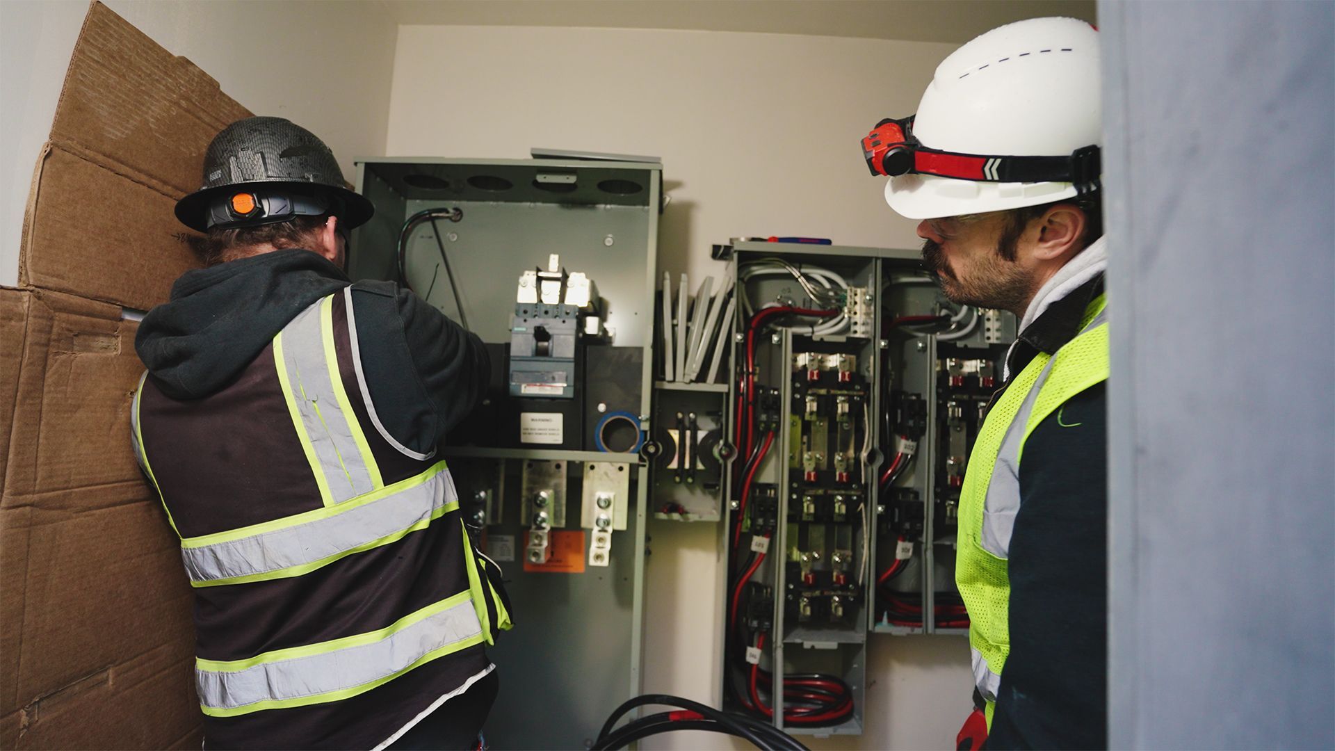 Two electricians in vests and hard hats work on electrical panel in a room