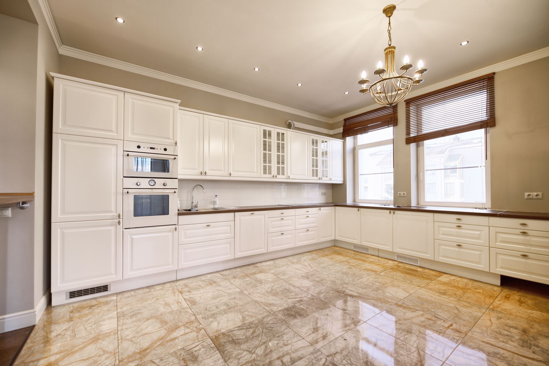 Bright white kitchen with marble floor, built-in ovens, chandelier, and large windows.