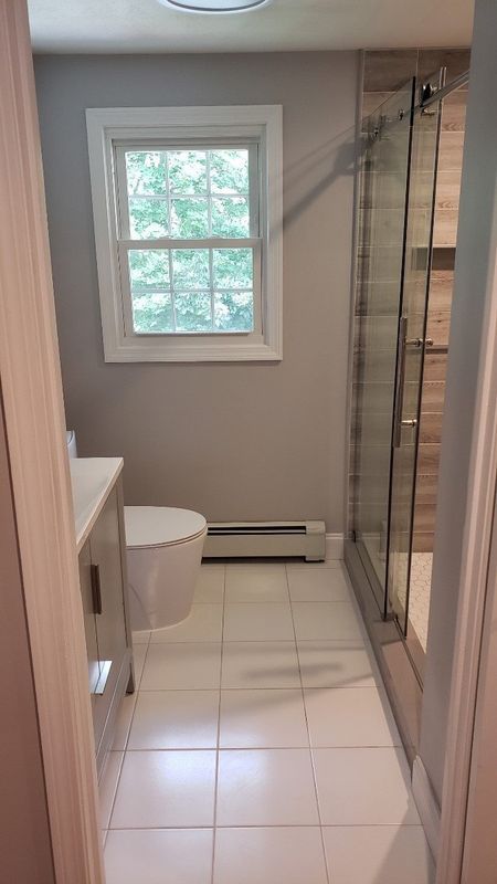 Small bathroom with white tile floor, toilet, window, and glass shower enclosure.