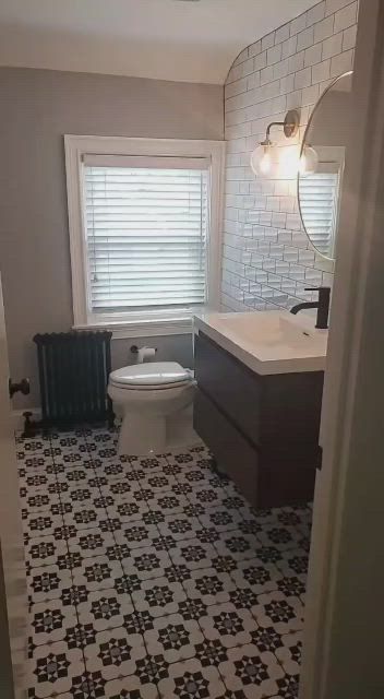 Small bathroom with patterned black-and-white floor, toilet, vanity, mirror, window blinds, and wall sconce.