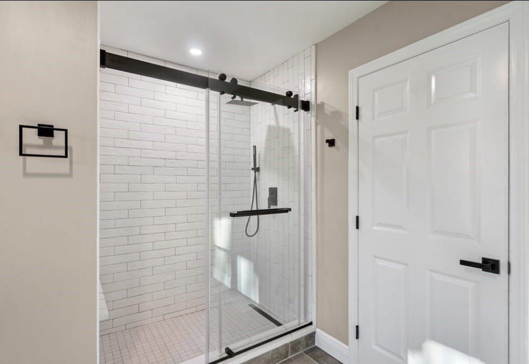 Modern bathroom shower with glass sliding door, white tile walls, and a white paneled door nearby