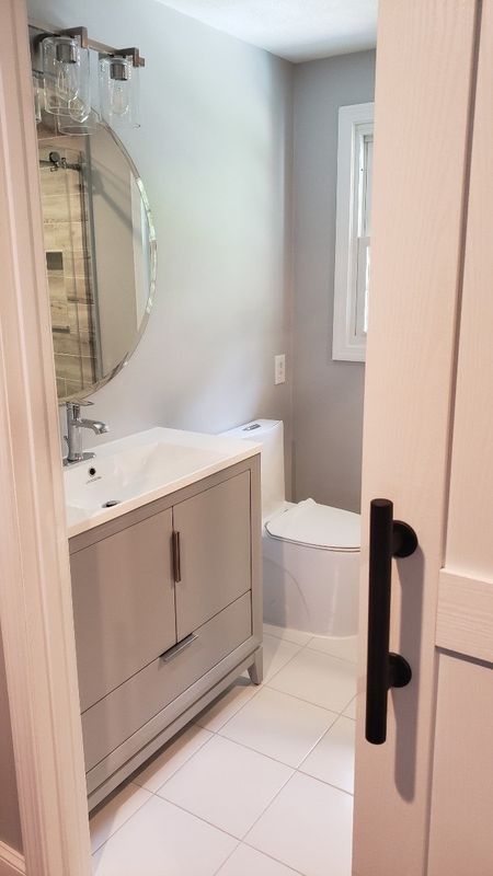 Narrow bathroom with white tile, vanity, round mirror, and toilet beside a door.