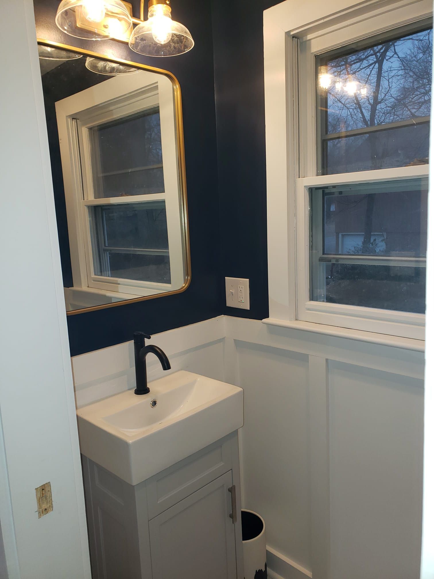 Small bathroom with navy walls, white wainscoting, pedestal sink, mirror, and window.