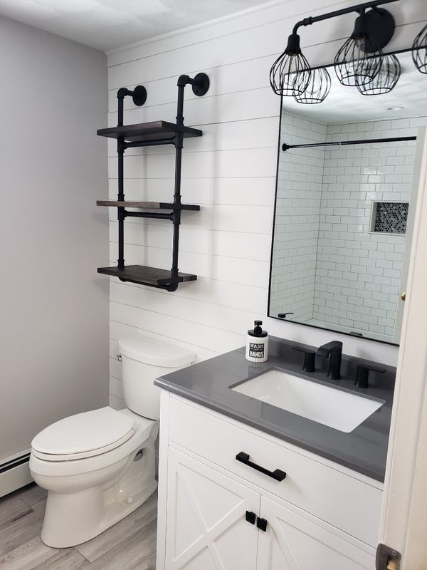 Small bathroom with toilet, vanity sink, mirror, and black wall shelves on white shiplap walls.