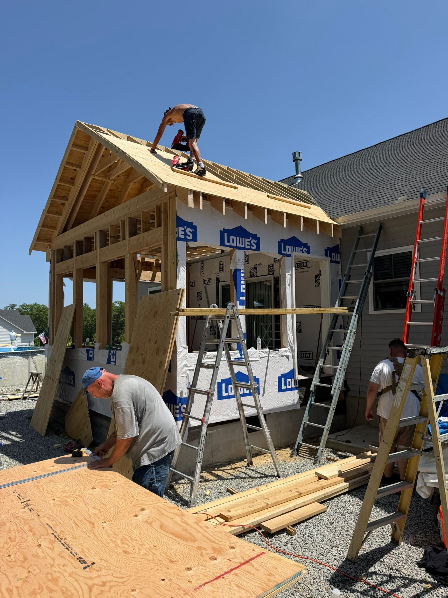 Workers building a house roof frame, with ladders, scaffolding, and exposed wood framing under a clear blue sky
