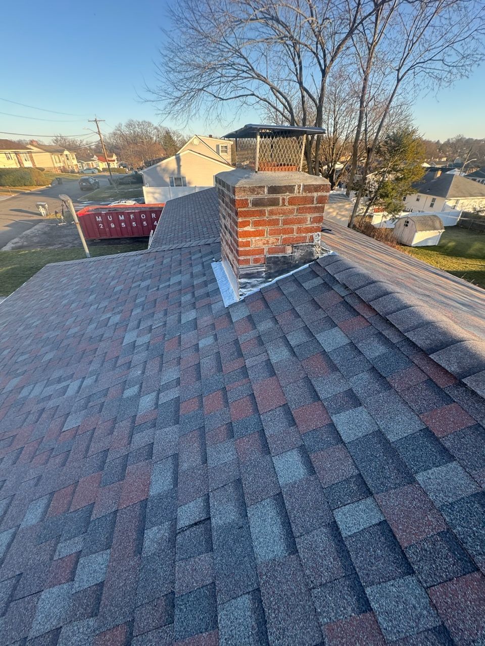 Rooftop with asphalt shingles and a brick chimney at sunset, trees and houses in the background