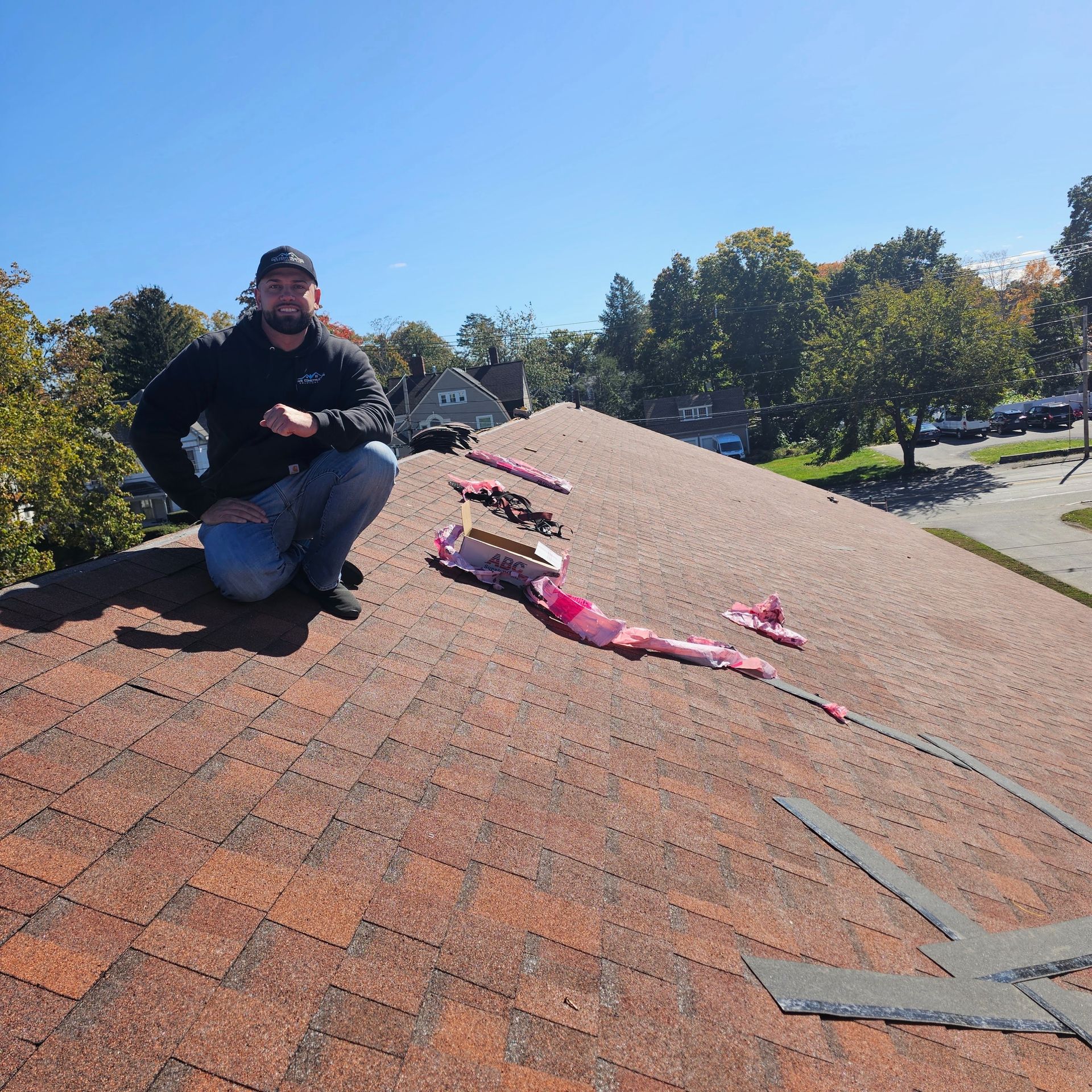Person kneeling on a shingled roof beside tools and pink straps under a clear blue sky