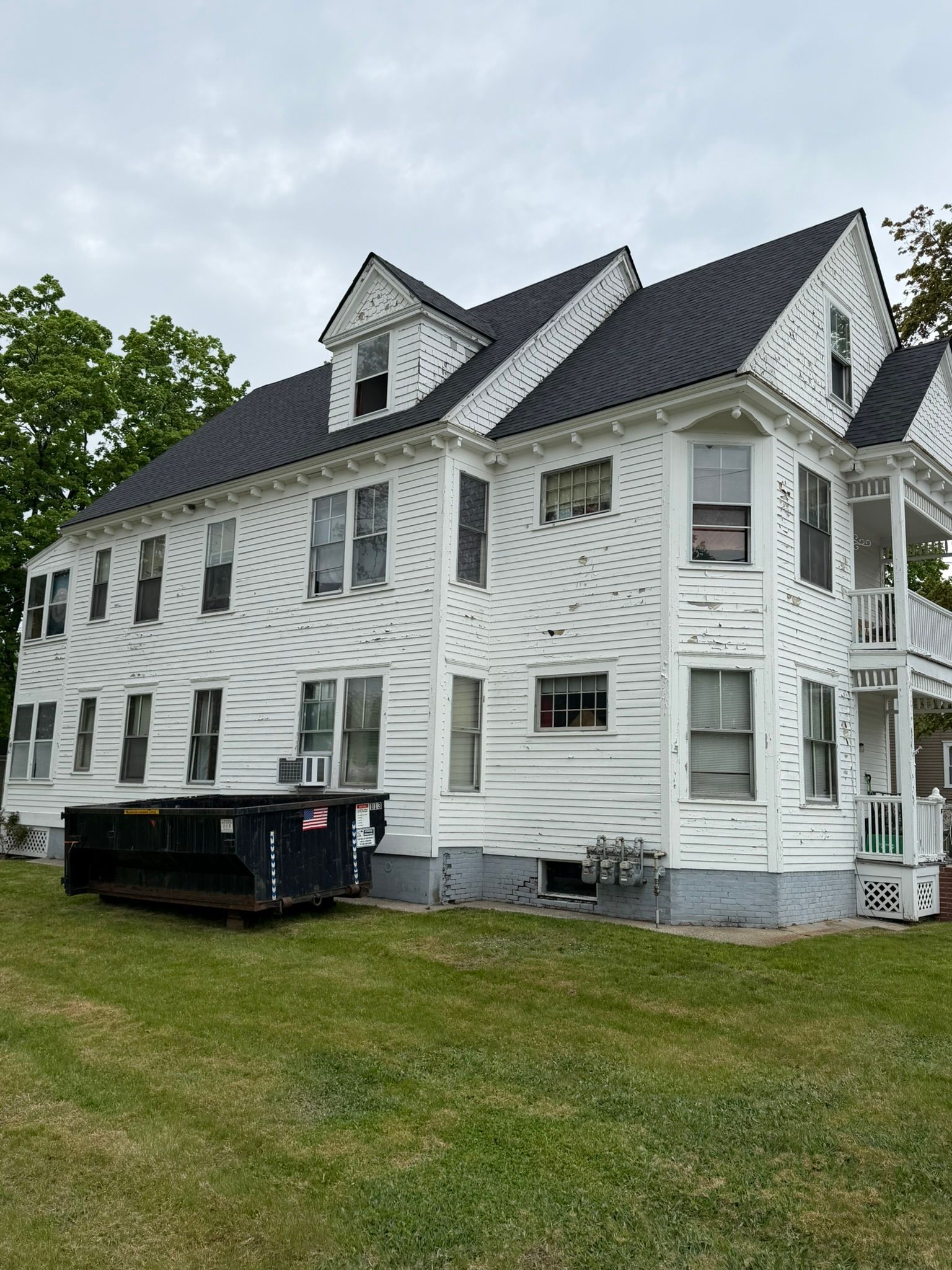 White three-story house with gray roof and black dumpster on a grassy lawn