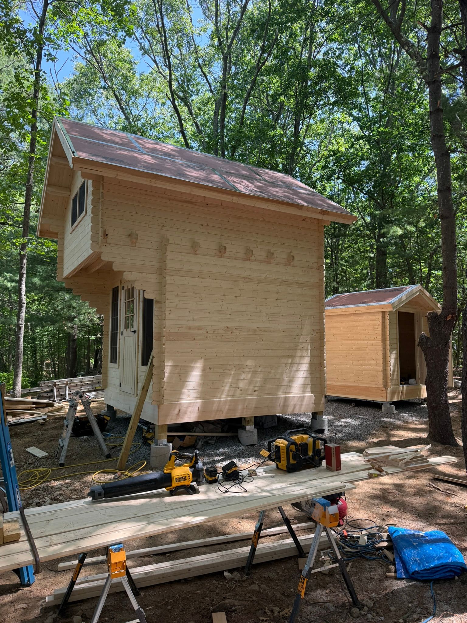 Two small unfinished wooden cabins under construction in a forest clearing, with tools and lumber on the ground