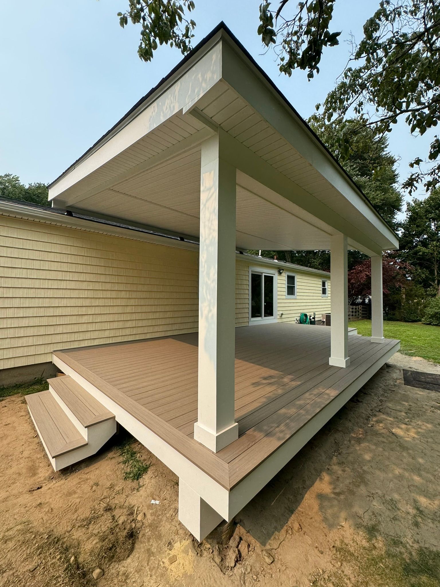 Front porch ramp with white posts and beige flooring beside a brick house under a roof overhang
