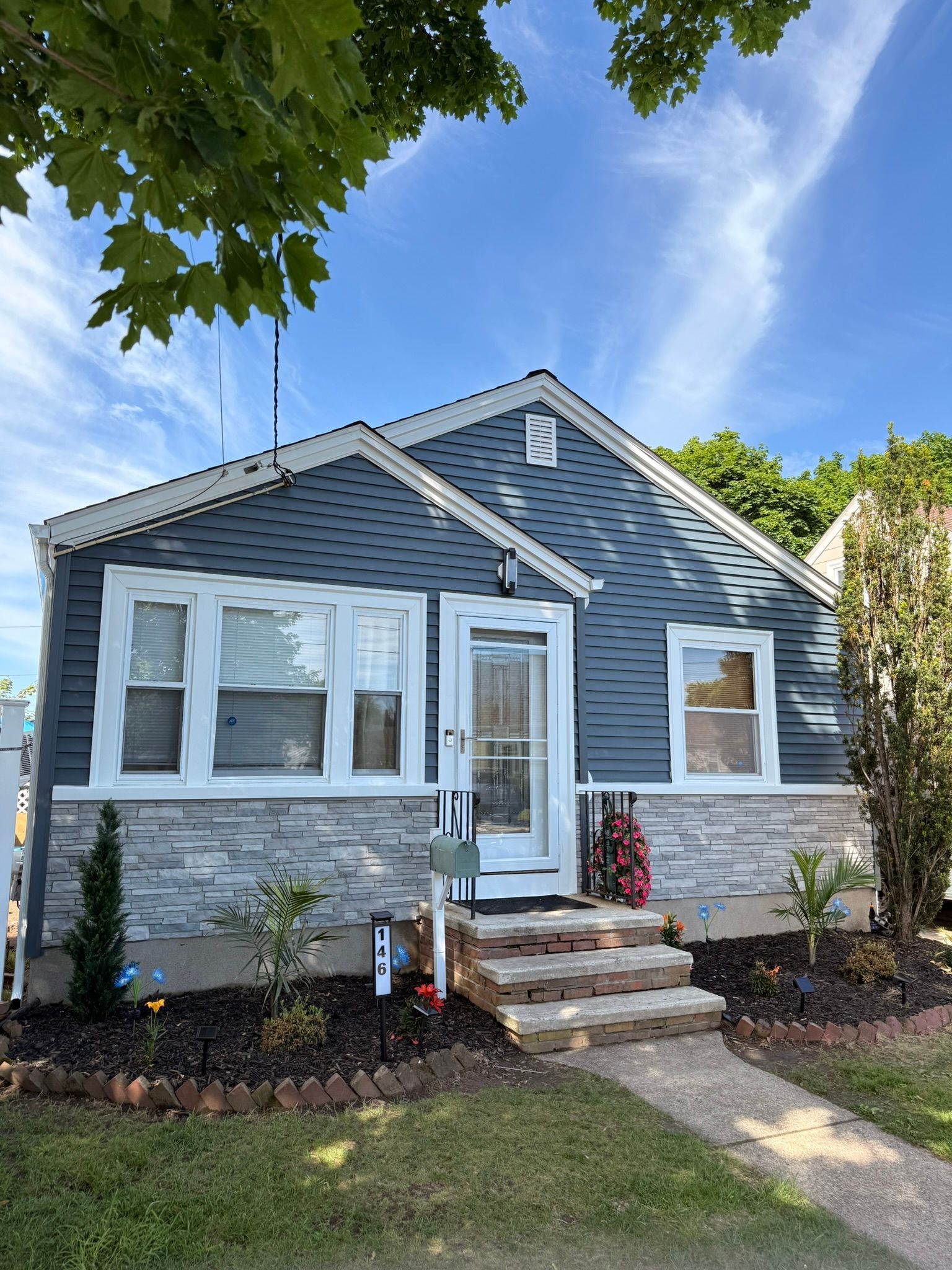 Small gray-blue house with white trim, front steps, and a tree-lined yard under a blue sky