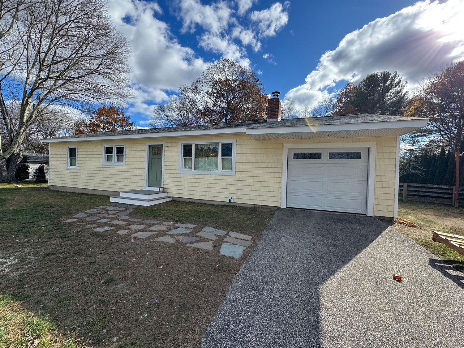 Single-story beige house with a garage, gravel driveway, and front steps under a cloudy blue sky