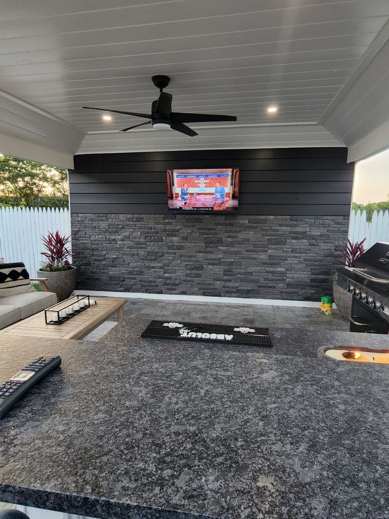 Covered outdoor patio with gray rug, ceiling fan, wall-mounted TV, and stone accent wall.