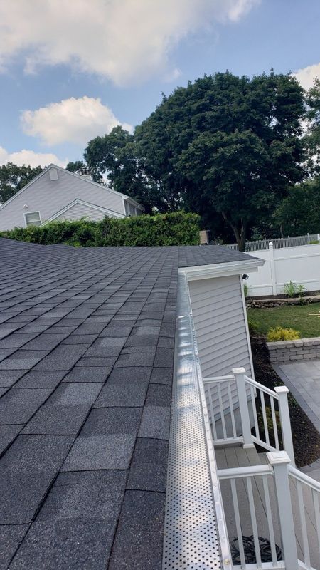Shingled roof beside white siding, railing, and a large tree under a partly cloudy sky.