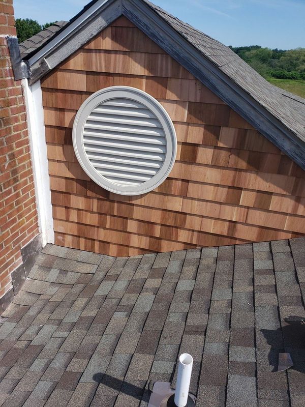 Rooftop with circular vent on shingled dormer above brick wall and chimney pipe.