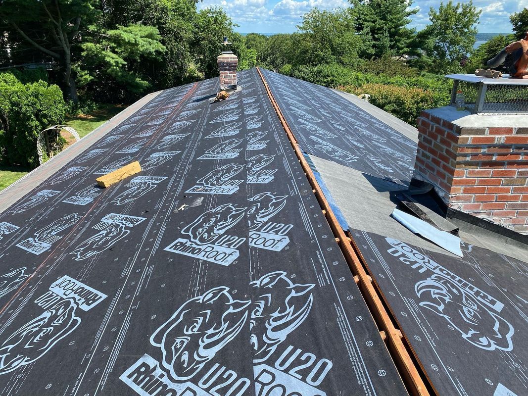 Partially installed roof underlayment on a sloped roof with brick chimney and trees nearby