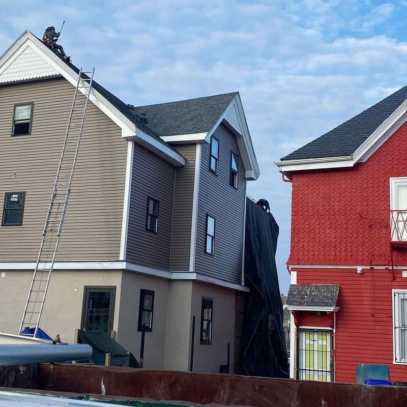 Two houses with beige and red siding, a ladder, and a tarp-covered structure between them under a cloudy sky