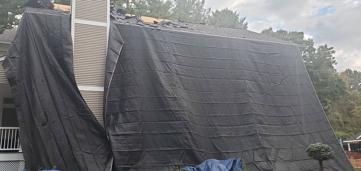 House roof covered in black tarps, with a chimney and trees in the background.