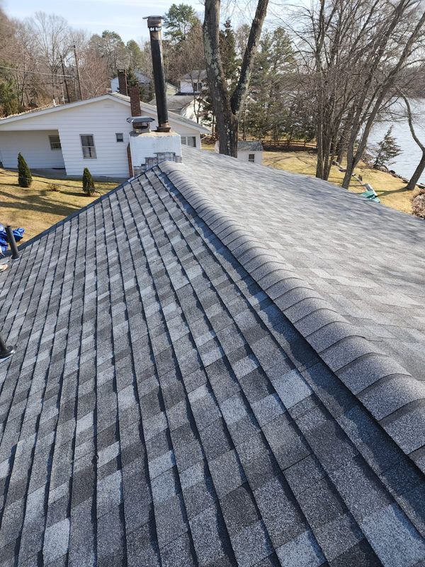 Roof shingles on a house, with dark new shingles in the foreground and an older gray roof section beside a chimney.