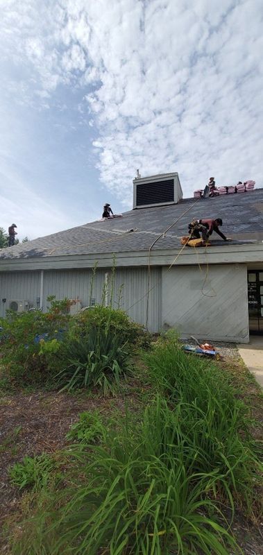 Workers on a sloped roof, replacing shingles on a house under a cloudy sky.