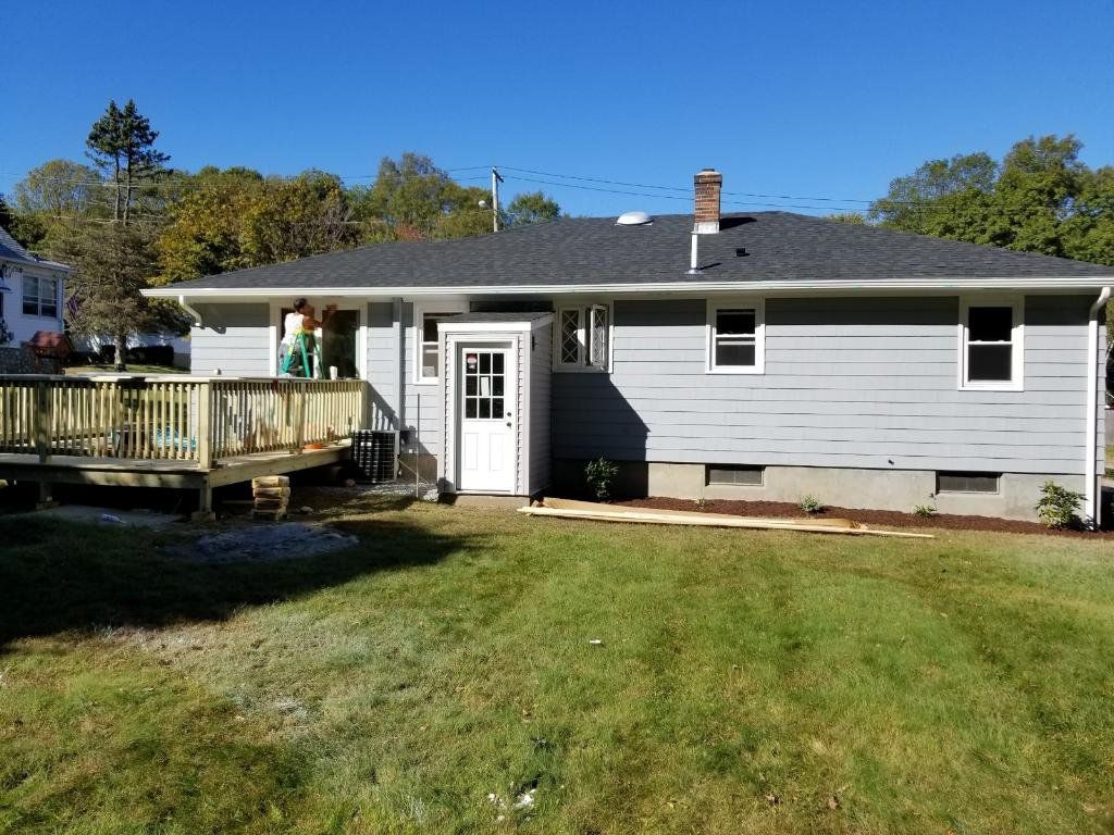 Single-story gray house with a wooden deck and front door, seen from a grassy backyard.