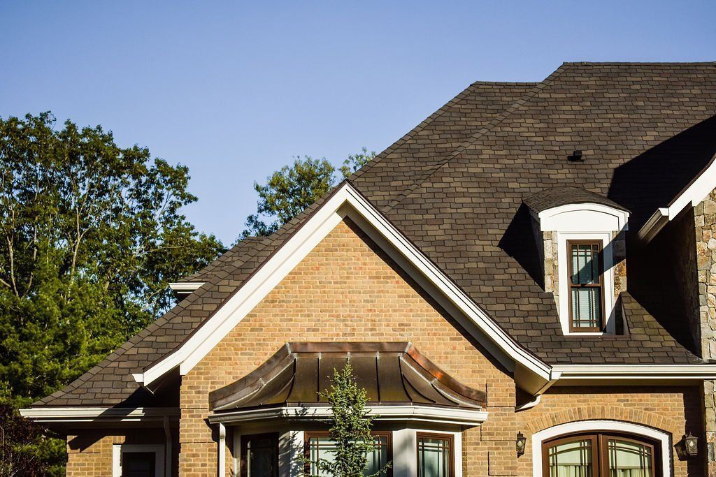Brick house with a steep shingled roof, white trim, and a tree at the left edge under a clear blue sky