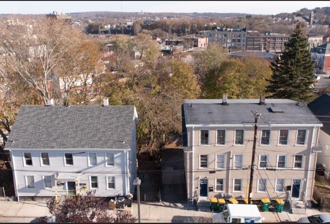 Two gray houses on a tree-lined street with a cityscape in the background
