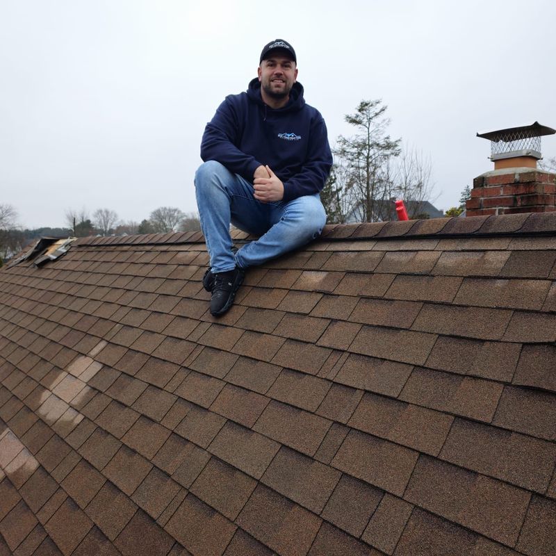 Person crouching on a shingled roof under an overcast sky, wearing a navy hoodie and jeans.