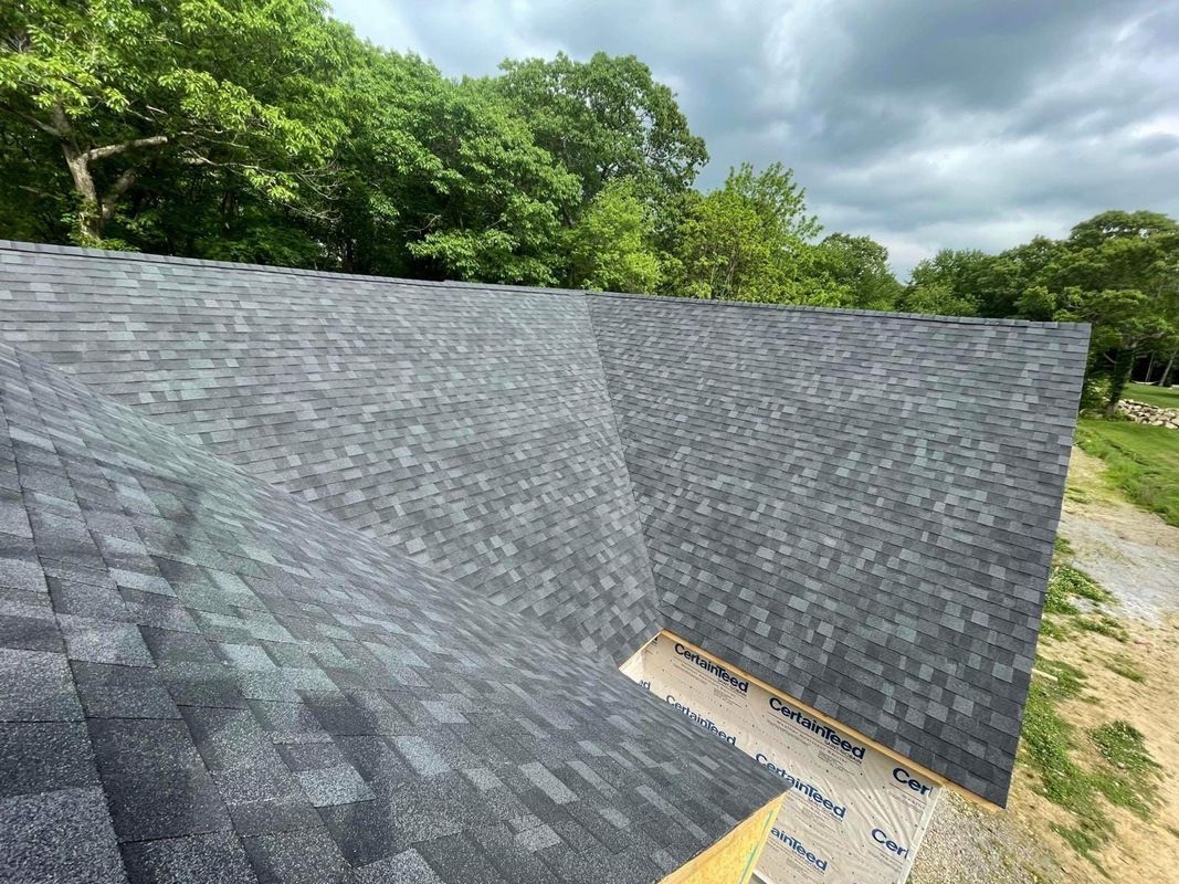 Roof covered in dark gray shingles against green trees and cloudy sky