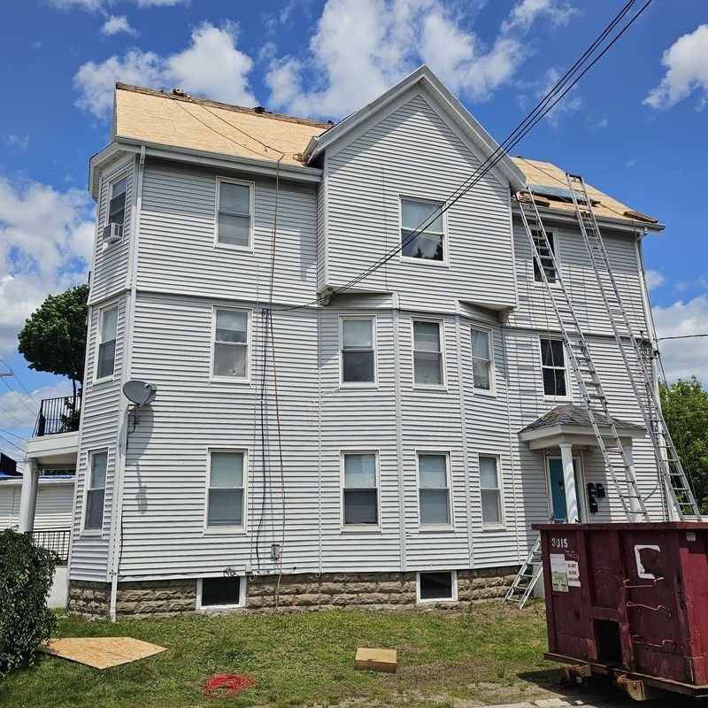 White multi-story house with damaged roof, a dumpster, and blue sky with clouds.