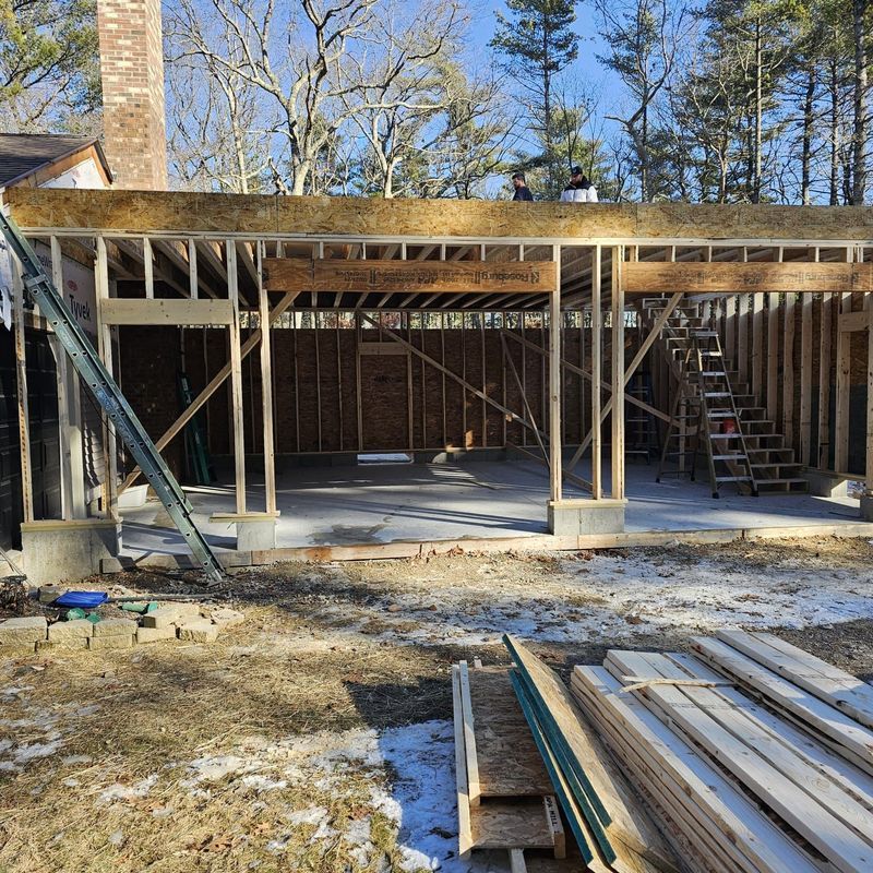 Partially built house frame with exposed lumber, concrete foundation, and stacked boards outside