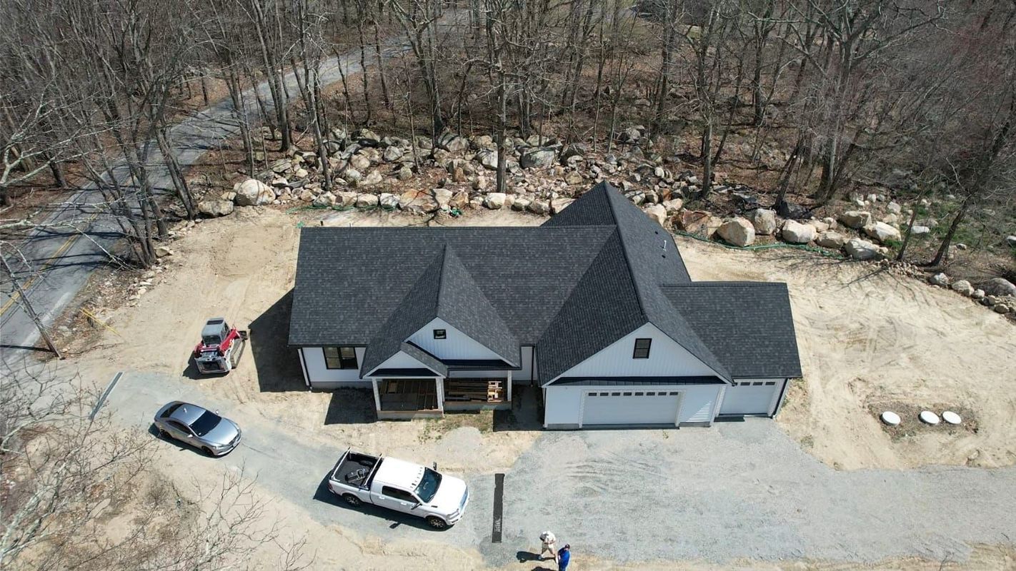 Aerial view of a gray house with a black roof, garage, and parked cars on a gravel lot near woods.