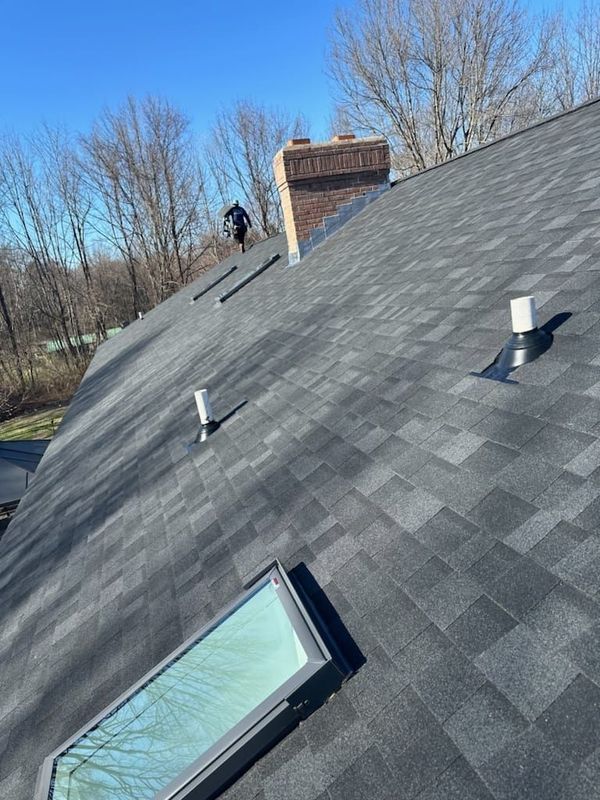 Sloped gray shingled roof with skylight, chimney, vents, and a worker near the ridge under a clear sky