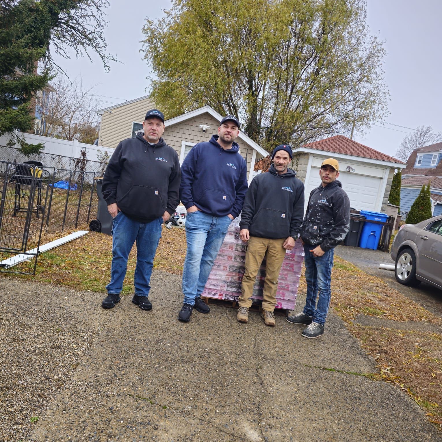 Four people posing on a driveway in front of a house with trees and a parked truck.