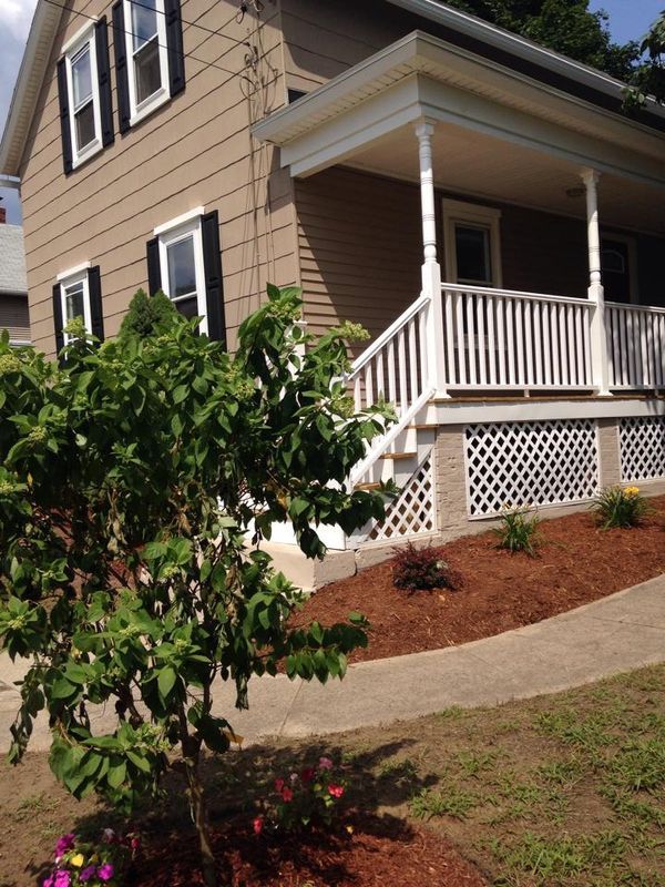 House porch with white railing, beige siding, and a small landscaped yard with shrubs and flowers