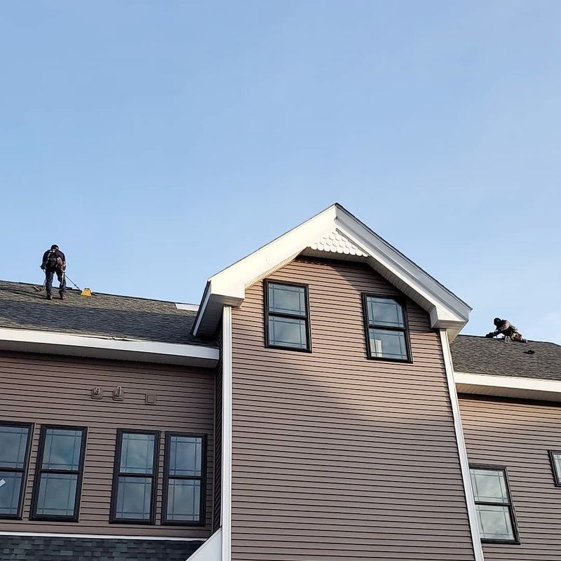 Two workers on a brown shingled roof of a house under a clear blue sky