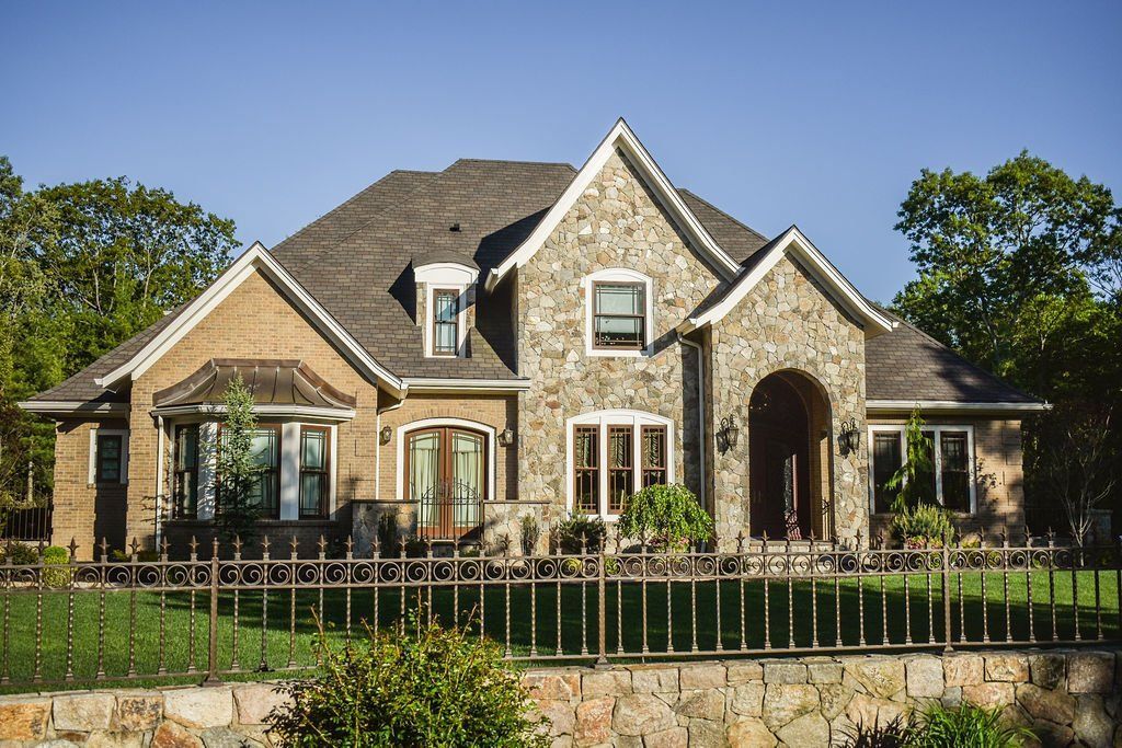 Stone house with a gabled roof, arched entry, and manicured front yard behind a low fence.