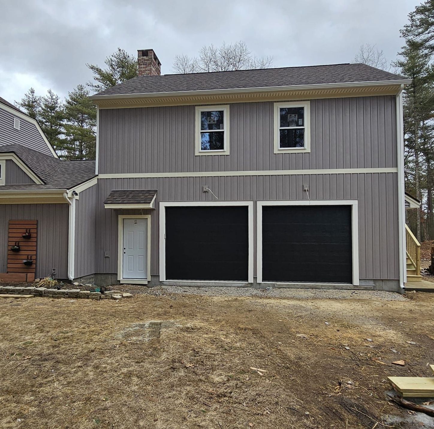 Two-story gray house with two black garage doors and a small side entrance, viewed from a bare yard