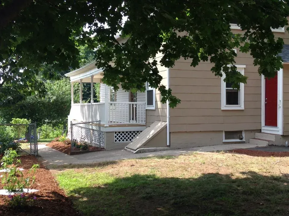 Small beige house with white porch, steps, and red front door under leafy trees.